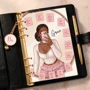 May include: A black leather planner with gold rings and a gold pen. The page features an illustration of a person in a white long-sleeve top and pink sweatpants holding a phone, surrounded by pink sticky notes with affirmations and the word Grace.