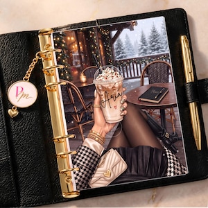 May include: A black leather planner with gold rings and a matching gold pen. The cover features an illustration of a person holding a coffee cup labeled Aria, wearing a houndstooth jacket, black skirt, and a small beige quilted handbag.