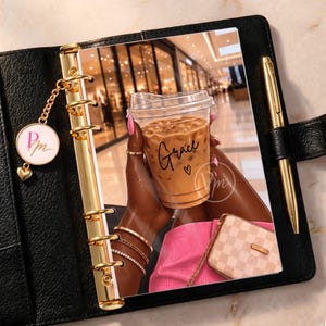 Könnte beinhalten: Ein schwarzer Lederplaner mit goldenen Ringen und einem goldenen Stift. Die Einlage zeigt eine Illustration von Händen, die einen Eiskaffee mit dem Namen Grace und einem Herz halten, dazu einen rosa Rock und eine karierte Handtasche.