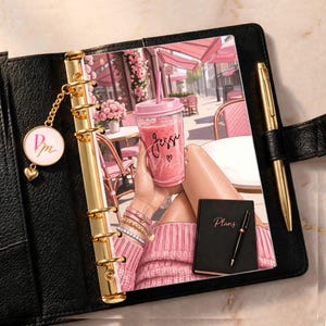 May include: A black leather binder with gold rings and a gold pen. The binder insert features an illustration of a person in a pink sweater holding a pink drink with the name Jessi and a heart. A small round charm with the letters PM hangs from the rings.