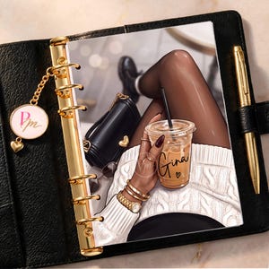 May include: A black leather planner with gold rings and a gold pen. The planner insert features an illustration of a person in a white cable-knit sweater holding an iced coffee cup labeled Gina, with a black handbag and a gold charm with PM text.