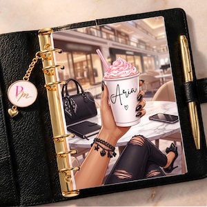 May include: A black leather planner with gold rings and a gold pen, featuring an insert illustration of a hand holding a white cup with the name Aria and a pink drink, a black handbag, and a smartphone on a marble table with a charm attached.