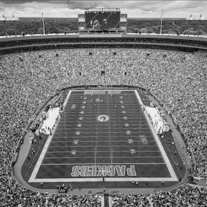 Puede incluir: Una vista panorámica en blanco y negro desde un ángulo elevado de un gran estadio de fútbol al aire libre lleno de espectadores. El campo rectangular presenta líneas de yardas, un logotipo G central y la palabra PACKERS en la zona de anotación.