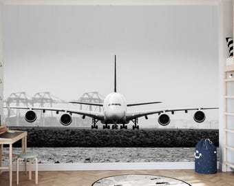 May include: Black and white photograph of a large passenger airplane on a runway, viewed head-on. The plane's wings extend horizontally, and its engines are visible. The background includes water, a rocky barrier, and industrial structures.