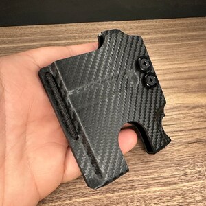 Puede incluir: Una cartera negra de fibra de carbono con una superficie texturizada y dos tornillos negros. La cartera tiene una ranura para efectivo y un bolsillo para tarjetas.