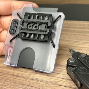 Puede incluir: Una cartera de pl&aacute;stico transparente con un porta tarjetas de metal negro y una correa negra con el texto "GGG" en ella. La cartera est&aacute; sobre una superficie de madera junto a una multiherramienta negra.