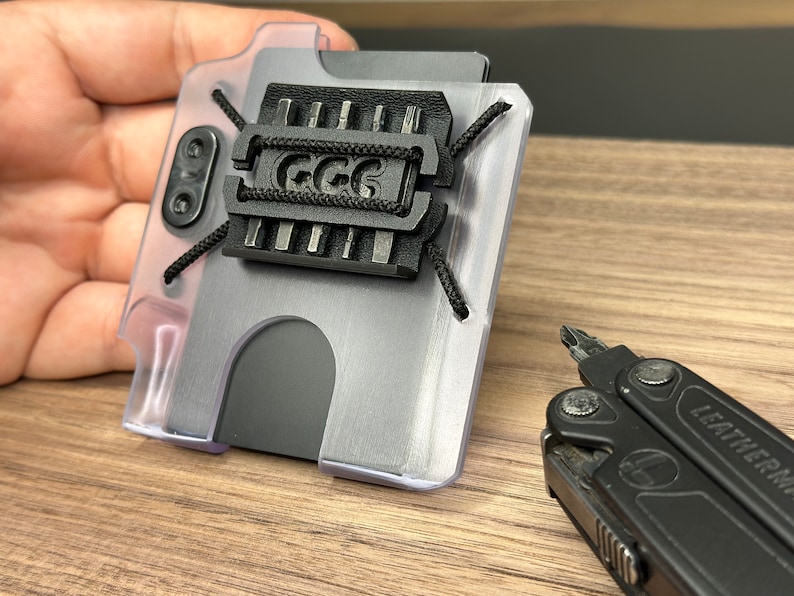 Puede incluir: Una cartera de pl&aacute;stico transparente con un porta tarjetas de metal negro y una correa negra con el texto "GGG" en ella. La cartera est&aacute; sobre una superficie de madera junto a una multiherramienta negra.