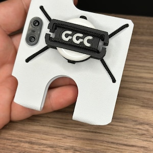 Puede incluir: Funda de tel&eacute;fono blanca con una correa negra y un c&iacute;rculo blanco con el texto "GCC" en negro.