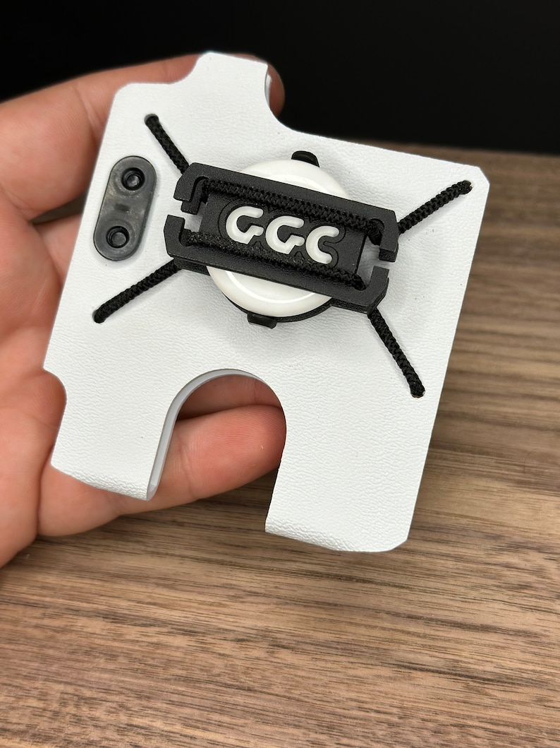 Puede incluir: Funda de tel&eacute;fono blanca con una correa negra y un c&iacute;rculo blanco con el texto "GCC" en negro.