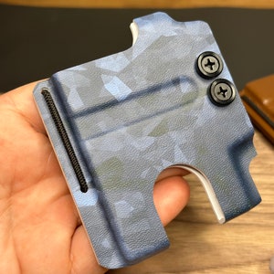 Puede incluir: Una cartera de cuero azul y blanco con estampado de camuflaje, una banda el&aacute;stica negra y dos tornillos negros. La cartera est&aacute; hecha de cuero y tiene una ranura para tarjetas.