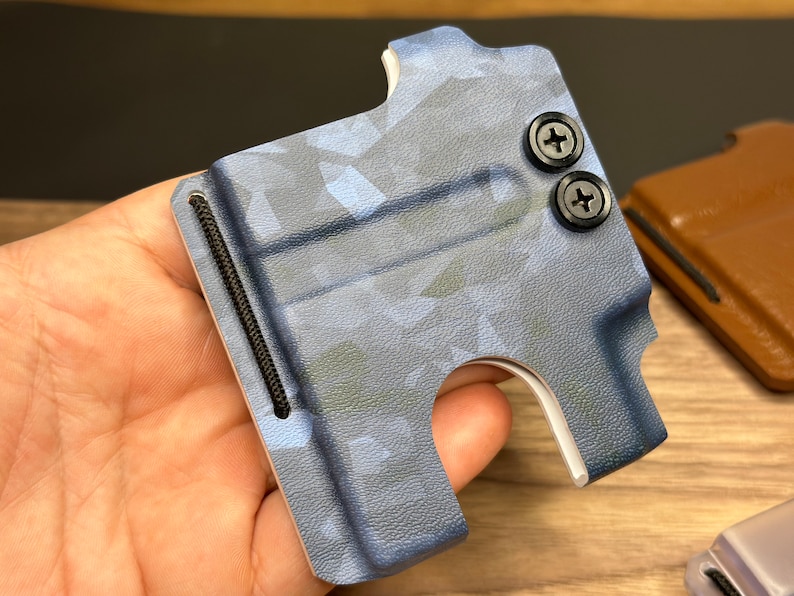 Puede incluir: Una cartera de cuero azul y blanco con estampado de camuflaje, una banda el&aacute;stica negra y dos tornillos negros. La cartera est&aacute; hecha de cuero y tiene una ranura para tarjetas.