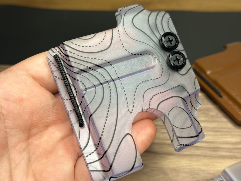 Puede incluir: Una cartera transparente con un dise&ntilde;o de mapa topogr&aacute;fico. La cartera tiene una banda el&aacute;stica negra y dos tornillos negros.
