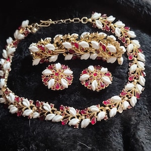 Op de afbeelding: Een vintage sieradenset met een ketting, armband en oorbellen. De set heeft een goudkleurige metalen basis met witte bladvormige accenten en rode edelsteen details. De oorbellen zijn rond met een bloemmotief.