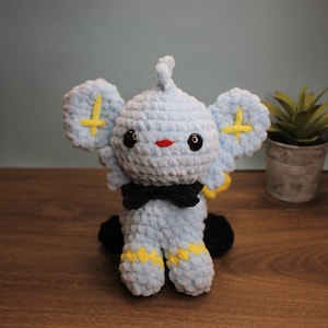 May include: A blue and yellow crocheted plush toy with black eyes and a black bow tie. The toy is sitting on a wooden surface.