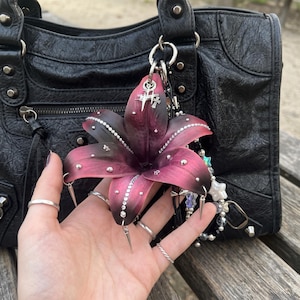May include: A dark purple and black lily flower charm featuring silver-toned metal spikes, clear rhinestones along the petals, and a small silver cross pendant. The charm is attached to a black textured leather handbag with silver studs.