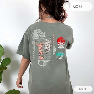 May include: A moss green t-shirt with a white graphic print of a Japanese demon mask, a pagoda, and a tree. The text "MOSS" is printed in white on the upper right corner of the image. The text "T-SHIRT" is printed in white on the lower right corner of the image.
