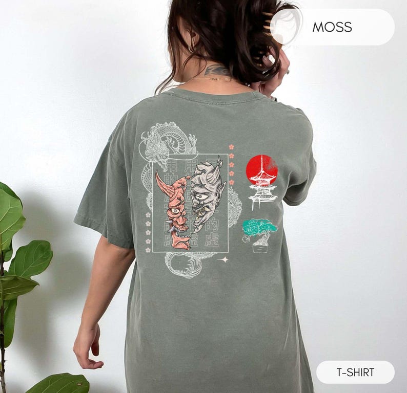 May include: A moss green t-shirt with a white graphic print of a Japanese demon mask, a pagoda, and a tree. The text "MOSS" is printed in white on the upper right corner of the image. The text "T-SHIRT" is printed in white on the lower right corner of the image.