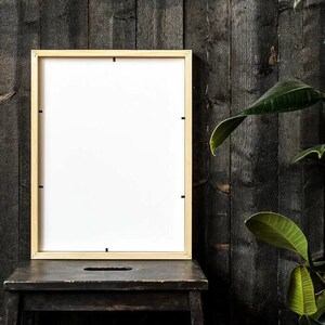 May include: A light wood picture frame with a white mat is sitting on a black wooden stool in front of a dark wood wall. A green plant is in the background.