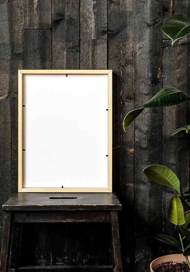 May include: A light wood picture frame with a white mat is sitting on a black wooden stool in front of a dark wood wall. A green plant is in the background.