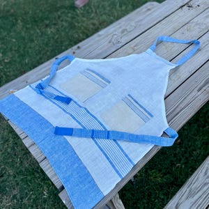Può includere: Un grembiule bianco con due tasche beige con accenti a righe blu, un laccio per il collo blu e lacci in vita blu. La parte inferiore del grembiule presenta un'ampia fascia blu con finitura testurizzata e ulteriori dettagli a righe blu.