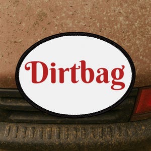 May include: Oval bumper sticker with the word "Dirtbag" in red, retro-style font. The sticker has a white background and a black border. The sticker is mounted on a brown, textured surface.