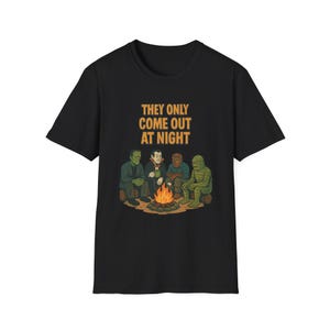 以下が含まれることがあります： 黒いTシャツ。フランケンシュタイン、ドラキュラ、狼男、黒いラグーンの生き物など、キャンプファイヤーの周りにいる古典的なモンスターのイラスト。テキスト「THEY ONLY COME OUT AT NIGHT」が上に印刷されています。
