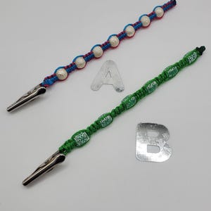 Puede incluir: Dos pinzas con cuentas. Una pinza es azul y roja con cuentas plateadas. La otra pinza es verde con cuentas estampadas verdes y blancas. Ambas pinzas tienen extremos de metal plateado. El fondo es blanco.