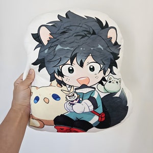 May include: A white plush pillow featuring a cartoon character with green hair and cat ears, wearing a teal and black outfit. The pillow also depicts two cat-like creatures and a small figure. The pillow is held by a person.