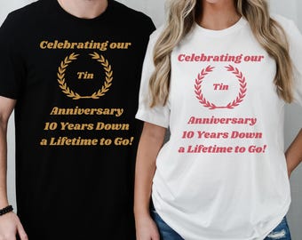 May include: Black and white t-shirts with the text "Celebrating our Tin Anniversary 10 Years Down a Lifetime to Go!" The text is in gold and red, with a laurel wreath design. The shirts are for a 10th anniversary.