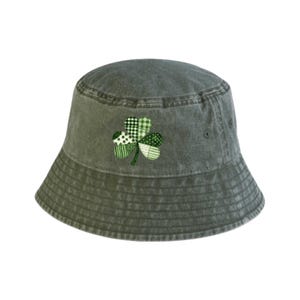 Puede incluir: Un sombrero de pescador verde con un trébol bordado. El trébol está hecho de varias telas verdes estampadas, incluyendo lunares, rayas y cuadros. El sombrero está hecho de una tela lavada y texturizada.