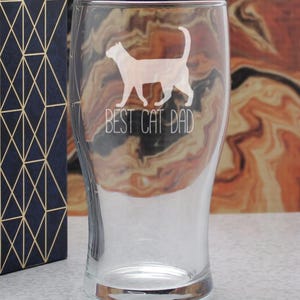 May include: A clear glass pint glass featuring an etched white silhouette of a cat standing in profile. Below the cat, the text BEST CAT DAD is etched in a clean, sans-serif font. The glass has a curved shape that tapers toward the base.