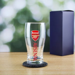 Może przedstawiać: Szklanka do piwa z przezroczystego szkła z logo klubu piłkarskiego Arsenal i napisem "ARSENAL" nadrukowanym pionowo na boku. Szklanka stoi na czarnym podkładce ze łupka.