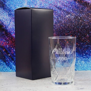 以下が含まれることがあります： ダイヤモンド模様の透明なガラスで、側面に「Phillipa's VODKA」という文字が刻まれています。ガラスは濃い青色の箱の隣にあります。