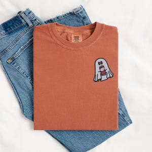 May include: A rust-colored t-shirt with a white ghost holding a red cup. The ghost has two black eyes and a black mouth. The t-shirt has a small tag on the bottom left corner that says "COMFORT COLORS".