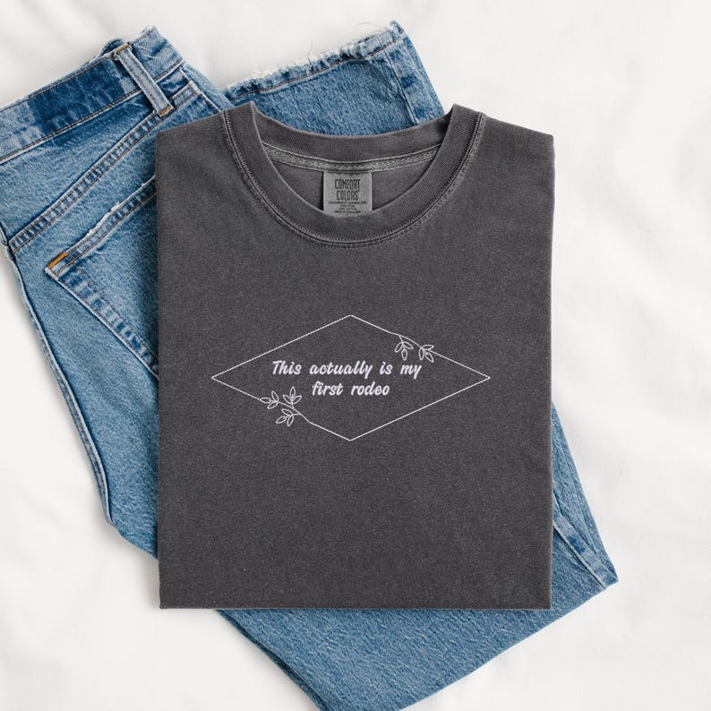 Pode incluir: Uma camiseta cinza escura com o texto "This actually is my first rodeo" em um diamante branco com detalhes florais.