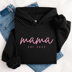 K&ouml;nnte beinhalten: Schwarzer Kapuzenpullover mit dem Text "mama est 2025" in rosa Schrift.
