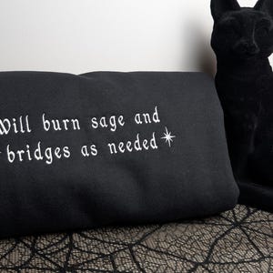 May include: A black sweatshirt featuring white embroidered text that reads "Will burn sage and bridges as needed" with star symbols on either side of the second line. A black cat figurine is positioned to the right of the folded garment.