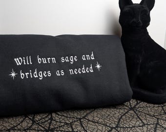 May include: A black sweatshirt featuring white embroidered text that reads "Will burn sage and bridges as needed" with star symbols on either side of the second line. A black cat figurine is positioned to the right of the folded garment.