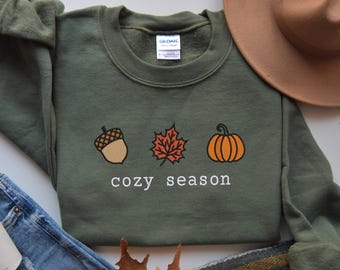 Op de afbeelding: Een groen sweatshirt met een witte afbeelding van een eikel, een esdoornblad en een pompoen. De tekst "cozy season" staat onder de afbeelding.