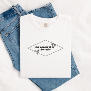 Pode incluir: Camiseta branca com um formato de diamante preto e o texto "This actually is my first rodeo" dentro do diamante. O diamante &eacute; contornado por uma linha preta e tem pequenos desenhos florais pretos de cada lado.
