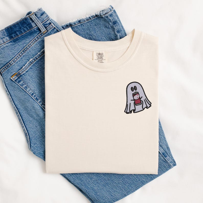 May include: A cream colored t-shirt with a ghost holding a red and white striped cup. The ghost is embroidered on the shirt. The shirt is folded and laid on top of a pair of blue jeans.