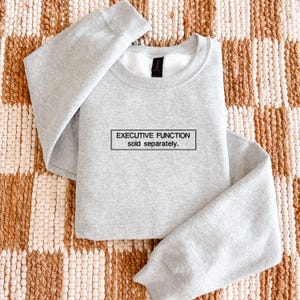 May include: A light gray crewneck sweatshirt featuring a black rectangular box on the chest. Inside the box, the text reads "EXECUTIVE FUNCTION sold separately." in a clean, sans-serif font. The garment has long sleeves and a ribbed collar.