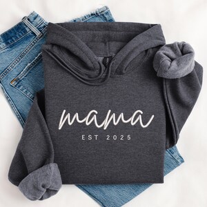 K&ouml;nnte beinhalten: Ein dunkelgrauer Kapuzenpullover mit dem Text "mama est 2025" in Wei&szlig;.