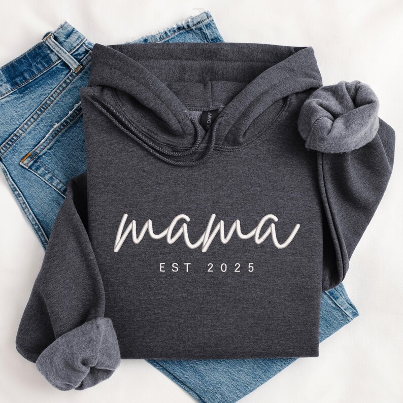 K&ouml;nnte beinhalten: Ein dunkelgrauer Kapuzenpullover mit dem Text "mama est 2025" in Wei&szlig;.