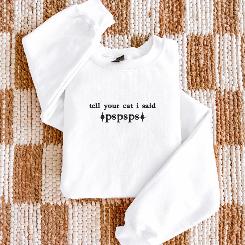 Pu&ograve; includere: Una felpa bianca a maniche lunghe con girocollo, caratterizzata da una scritta ricamata in nero sul petto che recita "tell your cat i said *pspsps*" con simboli a forma di stella, rifinita con polsini e scollo a costine.