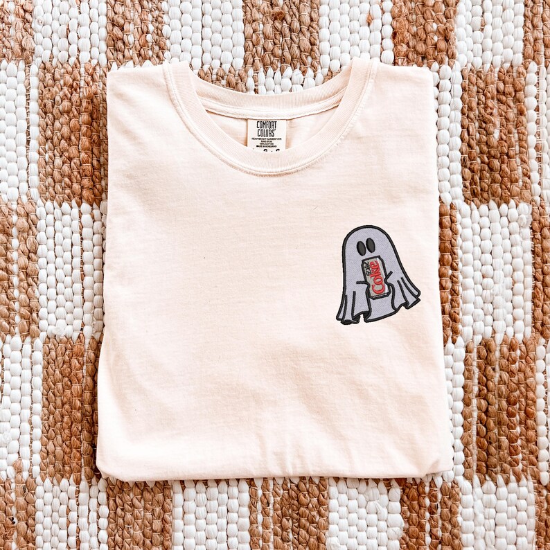 Op de afbeelding: Een licht perzikkleurig T-shirt met een ronde hals en een Comfort Colors merklabel. Op de linkerborst bevindt zich een geborduurde afbeelding van een grijze geest die een klein rood-wit frisdrankblikje vasthoudt met de tekst Coke.