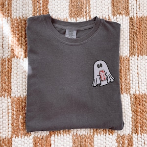 Op de afbeelding: Een antracietgrijs T-shirt met ronde hals en een kleine geborduurde afbeelding op de linkerborst van een wit spookje dat een rood Coca-Cola-blikje vasthoudt. Het binnenlabel vermeldt Comfort Colors, 100% katoen, made in Honduras.