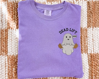 May include: A purple crew neck t-shirt featuring an embroidered graphic of a light gray ghost lifting a barbell. The text above the ghost reads DEAD-LIFT in black, and a small speech bubble next to the ghost contains the word BOO in black text.