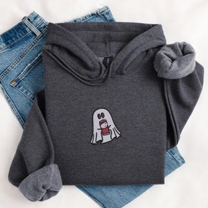 Könnte beinhalten: Ein dunkelgrauer Kapuzenpullover mit einem weißen Geist, der eine rote Dose mit dem Wort "Boo" hält. Der Geist ist auf der Vorderseite des Pullovers gestickt.