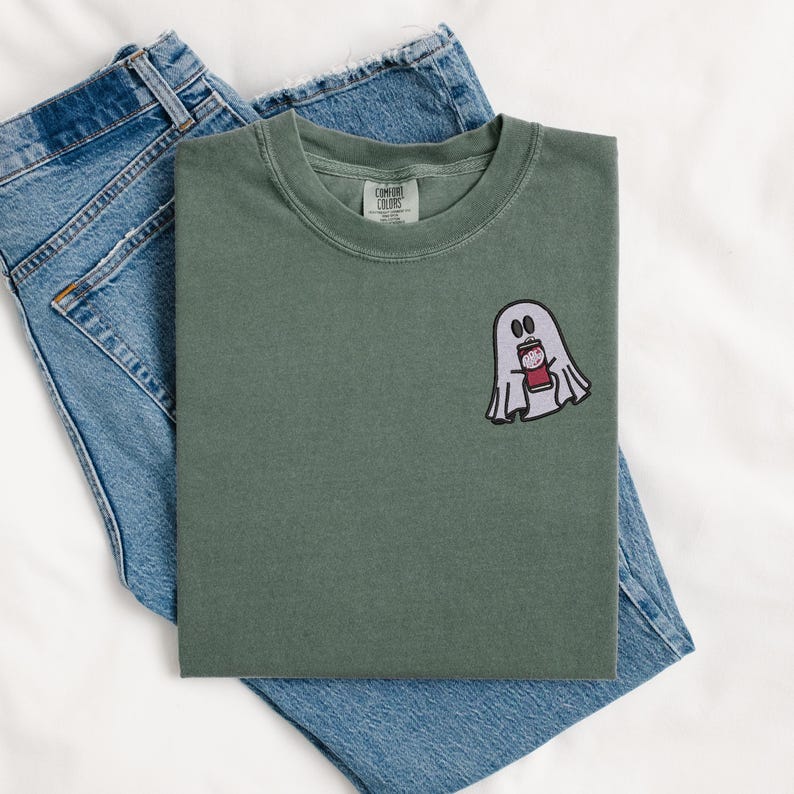 May include: A green t-shirt with a white ghost holding a red cup with a white design. The t-shirt has the text "COMFORT COLORS" on the tag.